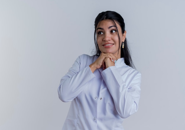 Souriante jeune femme médecin portant une robe médicale regardant le côté en gardant les mains ensemble sous le menton isolé