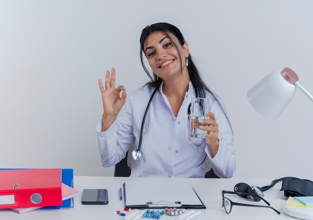Souriante jeune femme médecin portant une robe médicale et un stéthoscope assis au bureau avec des outils médicaux à la main tenant un verre d'eau faisant signe ok isolé