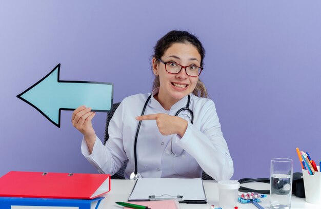 Souriante jeune femme médecin portant une robe médicale et un stéthoscope assis au bureau avec des outils médicaux à la marque de flèche tenant pointant vers le côté isolé