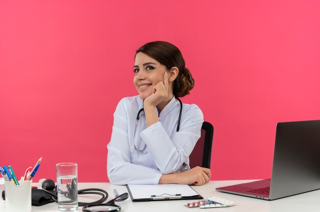 Souriante jeune femme médecin portant une robe médicale et un stéthoscope assis au bureau avec des outils médicaux et un ordinateur portable mettant les mains sur le bureau et sur le menton à la recherche tout droit isolé sur le mur rose