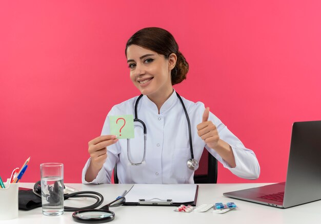 Souriante jeune femme médecin portant une robe médicale et un stéthoscope assis au bureau avec des outils médicaux et un ordinateur portable tenant un point d'interrogation montrant le pouce vers le haut isolé sur un mur rose