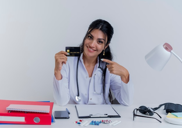 Souriante jeune femme médecin portant une robe médicale et un stéthoscope assis au bureau avec des outils médicaux à la recherche montrant la carte de crédit pointant vers elle isolé