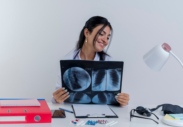 Souriante jeune femme médecin portant une robe médicale et un stéthoscope assis au bureau avec des outils médicaux tenant et regardant x-ray shot isolé