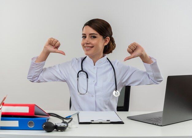 Souriante jeune femme médecin portant une robe médicale avec stéthoscope assis au bureau de travail sur ordinateur avec des outils médicaux pointe vers elle-même avec copie espace