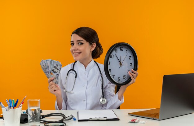 Souriante jeune femme médecin portant une robe médicale avec stéthoscope assis au bureau de travail sur ordinateur avec des outils médicaux tenant une horloge murale et de l'argent sur fond jaune d'isolement