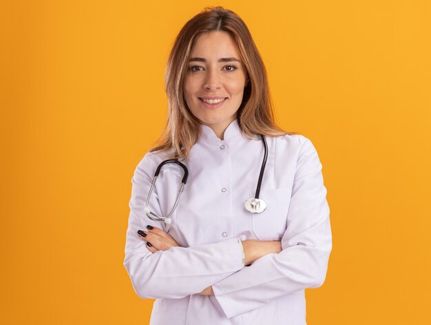 Souriante jeune femme médecin portant une robe médicale avec stéthoscope croisant les mains isolées sur le mur jaune