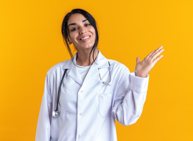 Souriante jeune femme médecin portant une robe médicale avec stéthoscope écartant la main isolée sur fond jaune