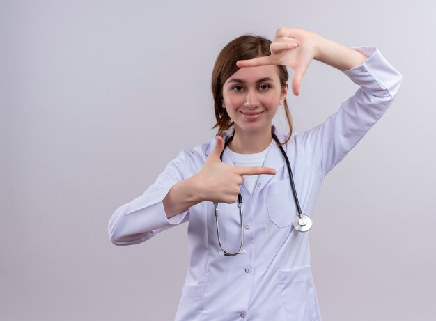 Souriante jeune femme médecin portant une robe médicale et un stéthoscope faisant un geste de cadre sur un mur blanc isolé