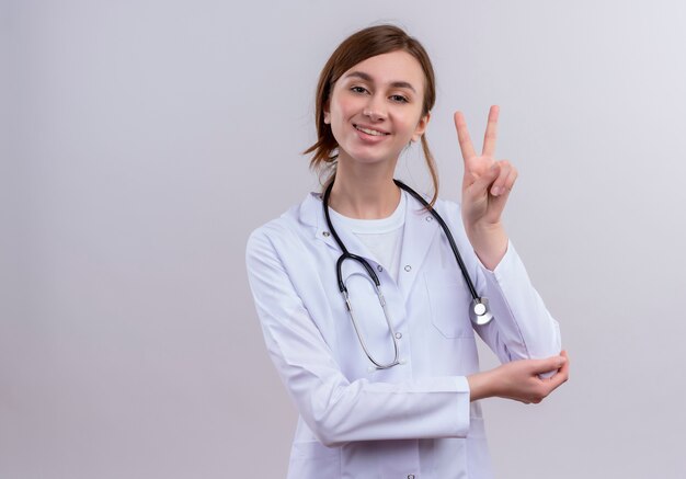Souriante jeune femme médecin portant une robe médicale et un stéthoscope faisant signe de paix sur un mur blanc isolé avec espace de copie
