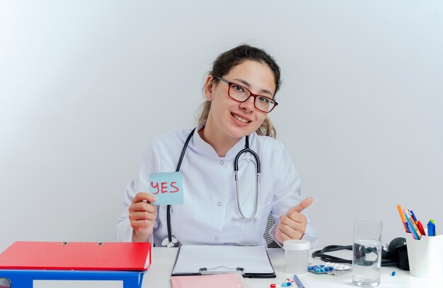 Souriante jeune femme médecin portant une robe médicale et un stéthoscope et des lunettes assis au bureau avec des outils médicaux à la tenue oui note montrant le pouce vers le haut isolé