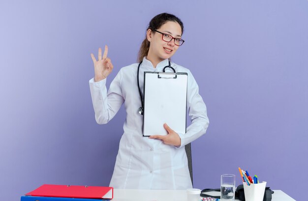 Souriante jeune femme médecin portant une robe médicale et un stéthoscope et des lunettes debout derrière un bureau avec des outils médicaux tenant le presse-papiers faisant signe ok à isolé