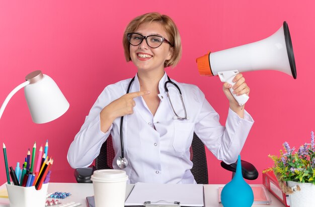 Souriante jeune femme médecin portant une robe médicale avec stéthoscope et lunettes est assise au bureau avec des outils médicaux tenant et pointe le haut-parleur isolé sur fond rose