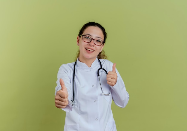 Souriante jeune femme médecin portant une robe médicale et un stéthoscope avec des lunettes montrant les pouces vers le haut isolé