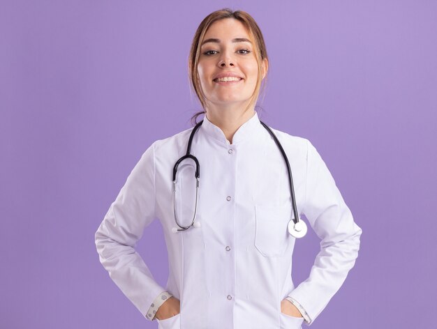 Souriante jeune femme médecin portant une robe médicale avec stéthoscope mettant les mains sur la poche isolée sur le mur violet