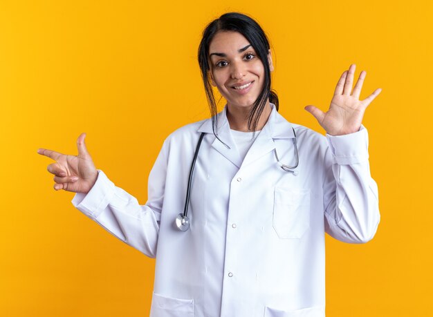 Souriante jeune femme médecin portant une robe médicale avec stéthoscope montrant différents numéros isolés sur fond jaune