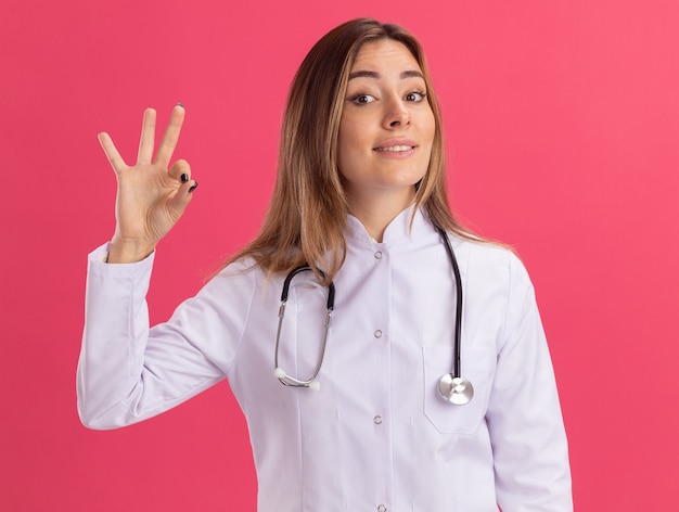 Souriante jeune femme médecin portant une robe médicale avec stéthoscope montrant un geste correct isolé sur un mur rose