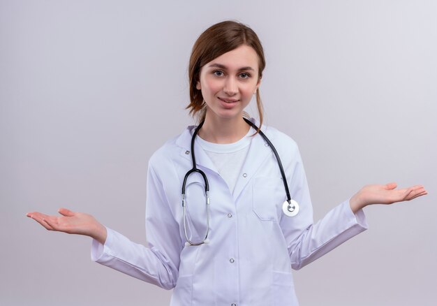 Souriante jeune femme médecin portant une robe médicale et un stéthoscope montrant des mains vides sur un mur blanc isolé