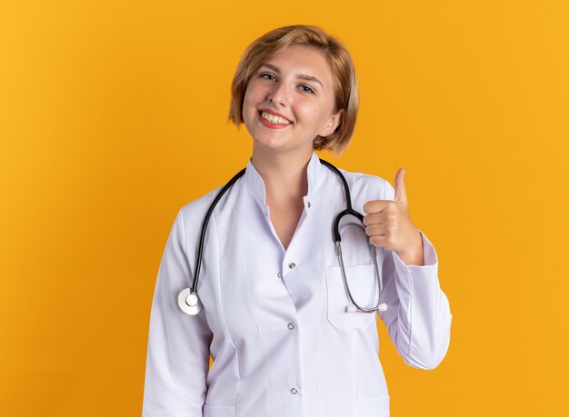 Souriante jeune femme médecin portant une robe médicale avec stéthoscope montrant le pouce vers le haut isolé sur fond orange
