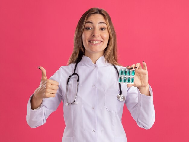 Souriante jeune femme médecin portant une robe médicale avec stéthoscope tenant des pilules montrant le pouce vers le haut isolé sur un mur rose