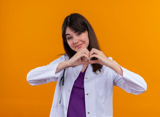 Souriante jeune femme médecin en robe médicale avec coeur de gestes stéthoscope avec les deux mains sur fond orange isolé avec espace copie