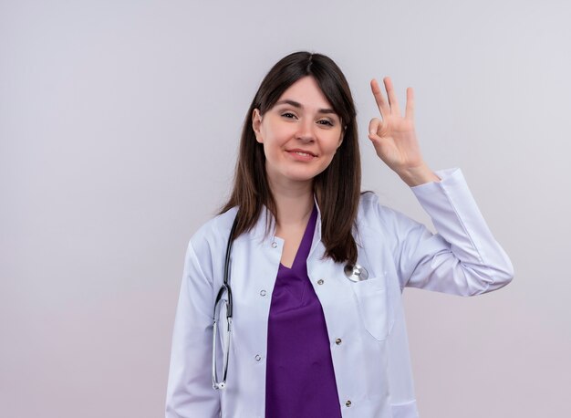 Souriante jeune femme médecin en robe médicale avec des gestes de stéthoscope ok sur fond blanc isolé avec copie espace