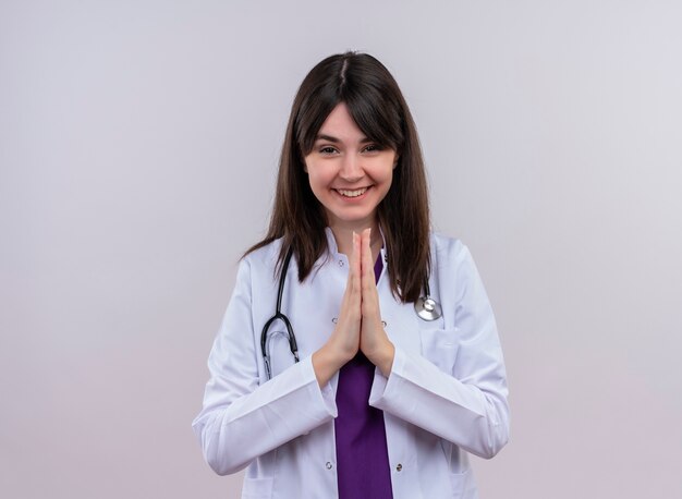 Souriante jeune femme médecin en robe médicale avec stéthoscope tient les mains ensemble sur fond blanc isolé avec espace copie