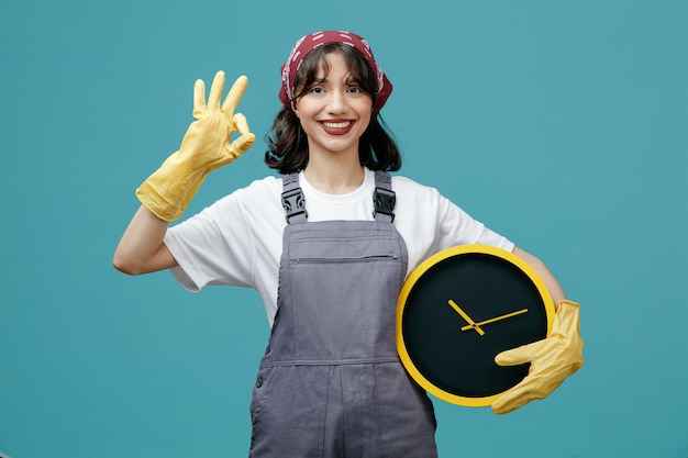 Souriante jeune femme nettoyante portant un bandana uniforme et des gants en caoutchouc tenant une horloge regardant la caméra montrant un signe ok isolé sur fond bleu