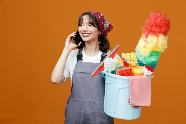 Souriante jeune femme nettoyante portant l'uniforme et le bandana tenant un seau d'outils de nettoyage regardant à côté tout en parlant au téléphone isolé sur fond orange