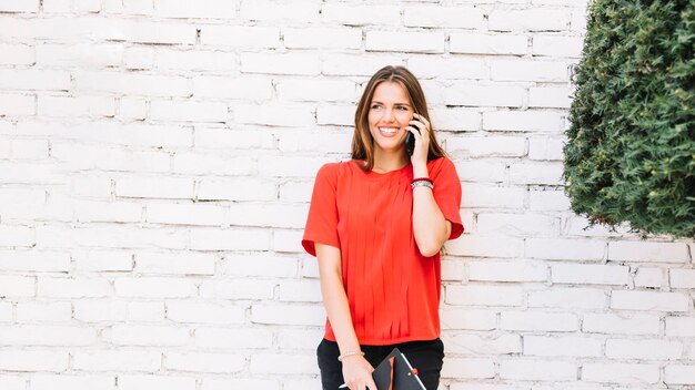 Souriante jeune femme parlant sur un smartphone devant un mur de briques