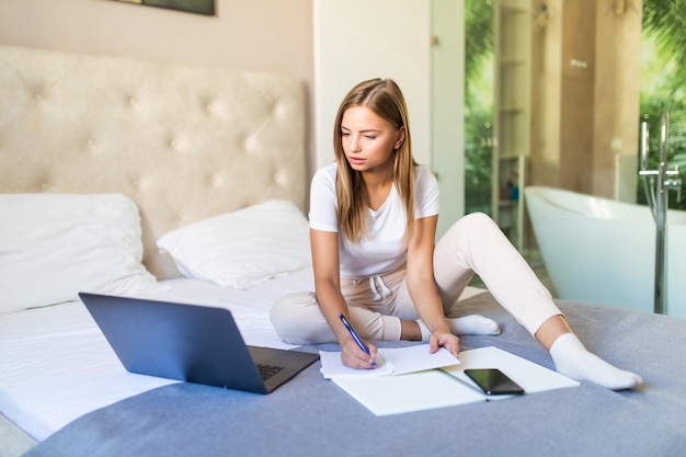 Souriante jeune femme payant ses factures en ligne avec un ordinateur portable dans la chambre à la maison