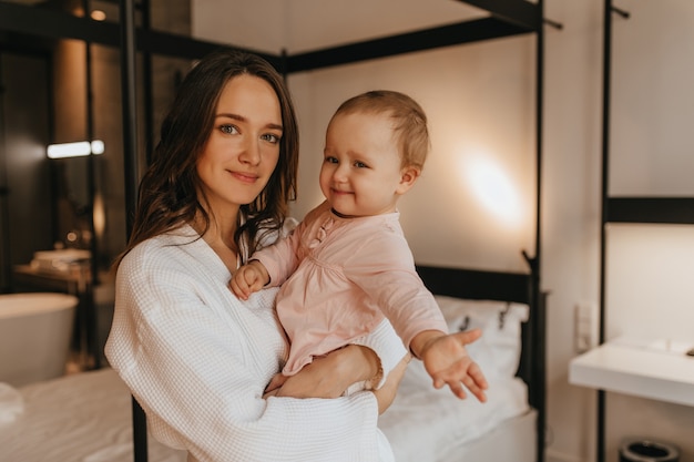 Souriante jeune femme en peignoir et sa petite fille mignonne souriant et posant dans la chambre.