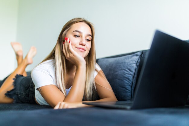 Souriante jeune femme portant sur un canapé et utilisant un ordinateur portable à la maison