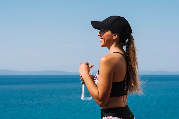 Souriante jeune femme portant casquette debout devant la mer, tenant une bouteille d&#39;eau à la main