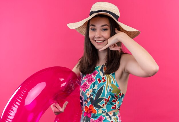 Souriante jeune femme portant un chapeau tenant l'anneau de bain et faisant signe de téléphone sur un mur rose isolé