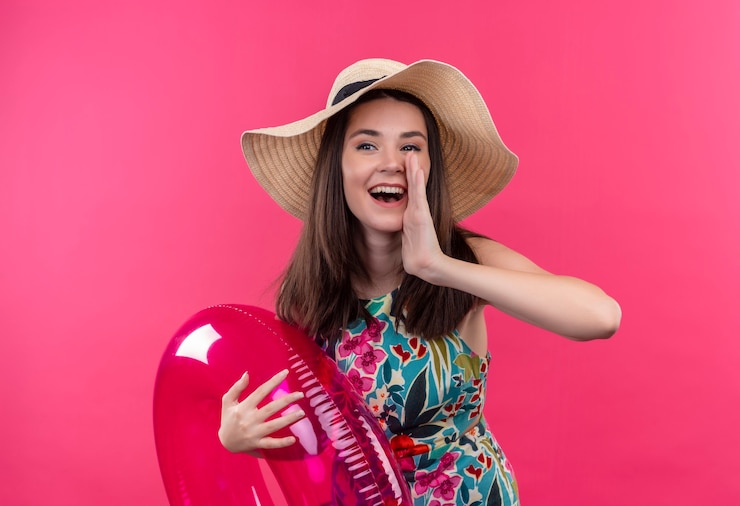Photo gratuit souriante jeune femme portant un chapeau tenant l'anneau de bain et tenant la main près de la bouche sur un mur rose isolé