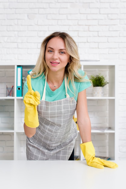 Photo gratuite souriante jeune femme portant un tablier et un gant jaune pointant l'index en regardant la caméra