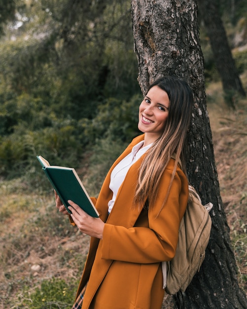 Souriante jeune femme posant sous l'arbre avec un livre à la main