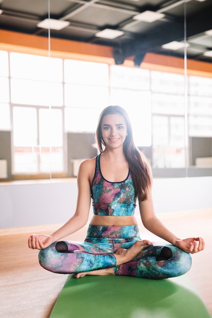 Souriante jeune femme en posture de lotus en regardant la caméra assis sur un tapis de yoga