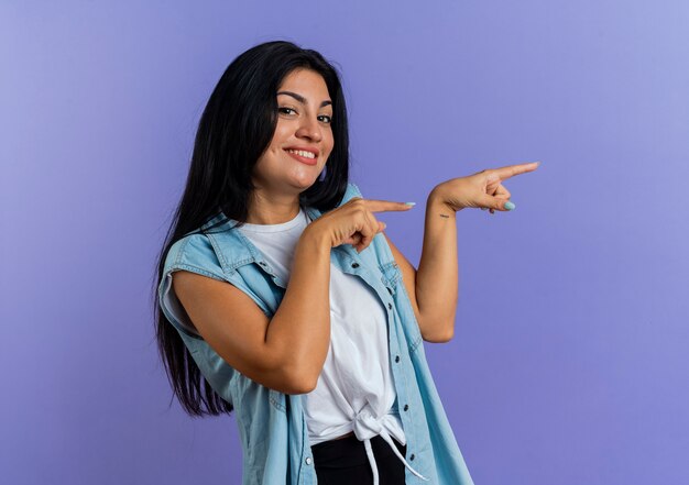 Souriante jeune femme de race blanche pointe sur le côté avec deux mains regardant la caméra isolée sur fond violet avec espace copie