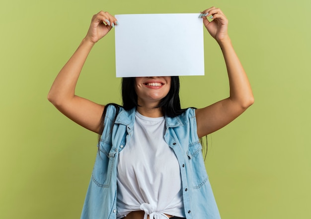 Photo gratuite souriante jeune femme de race blanche tient une feuille de papier au-dessus de la tête en fermant les yeux