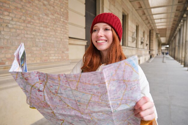 Photo gratuite souriante jeune femme rousse au chapeau rouge regarde la carte papier pour rechercher le tourisme d'attraction touristique et