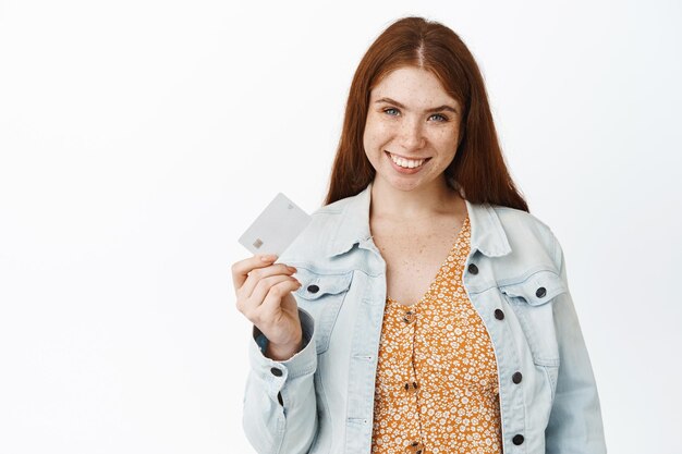 Souriante jeune femme rousse montrant une carte de crédit recommandant un magasin ou une banque fond blanc