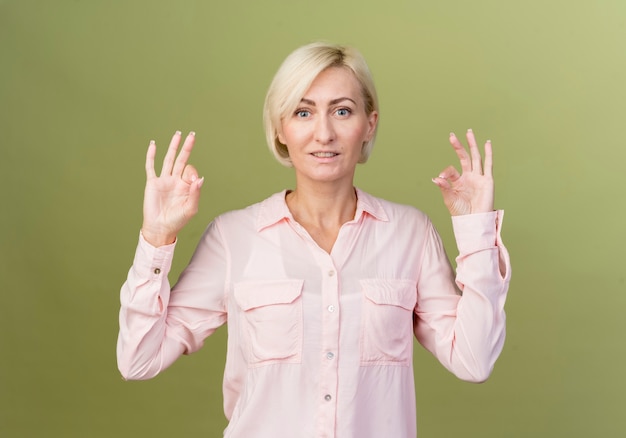 Souriante jeune femme slave blonde montrant le geste de paix isolé sur mur vert olive