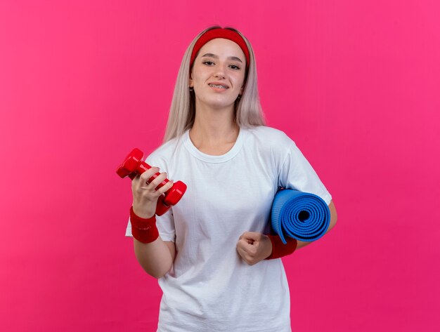 Souriante jeune femme sportive avec des accolades portant un bandeau et des bracelets tient un tapis de sport et un haltère isolé sur un mur rose