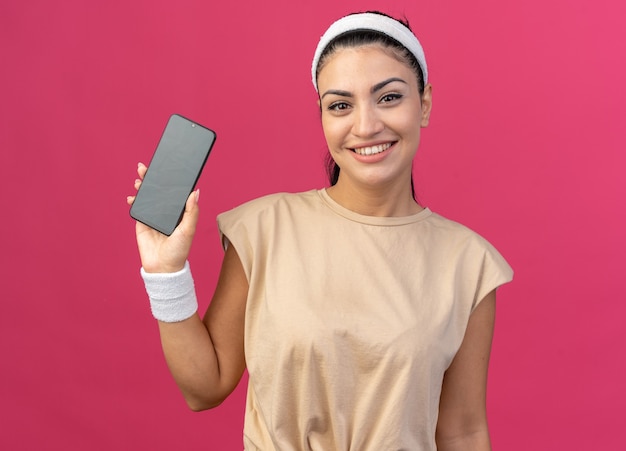 Souriante jeune femme sportive caucasienne portant un bandeau et des bracelets regardant à l'avant montrant un téléphone portable à une caméra isolée sur un mur rose