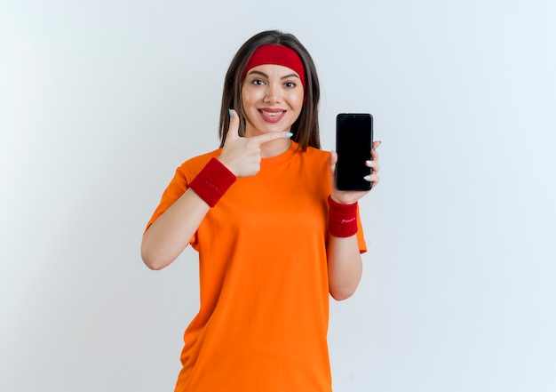 Souriante jeune femme sportive portant un bandeau et des bracelets montrant un téléphone mobile pointant vers elle à la recherche d'isolement