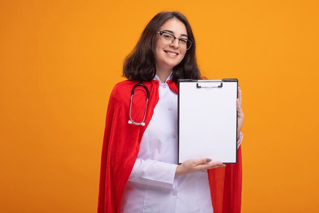 Souriante jeune femme super-héros en cape rouge portant un uniforme de médecin et un stéthoscope avec des lunettes montrant le presse-papiers à l'avant regardant à l'avant isolé sur un mur orange