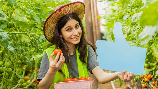 Souriante jeune femme tenant un panneau bleu et une tomate mûre à la serre