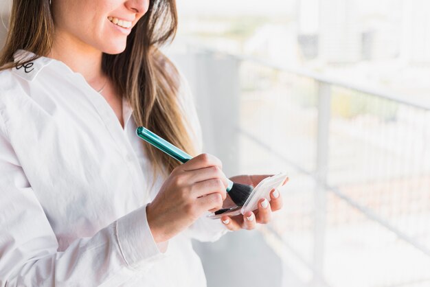 Souriante jeune femme tenant le pinceau à maquillage et la poudre compacte