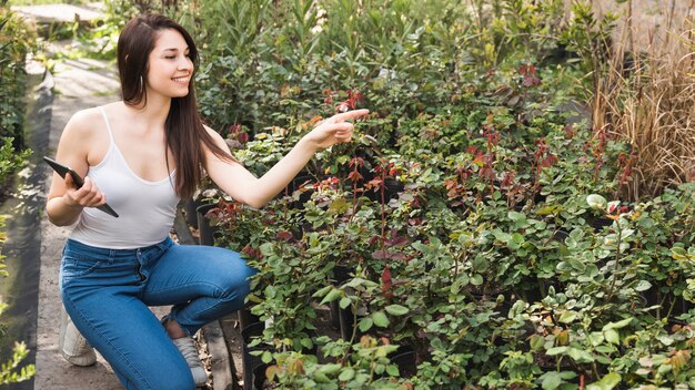 Souriante jeune femme tenant une tablette numérique dans la main, pointant vers les plantes dans le jardin